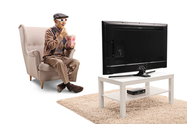 Elderly man seated in an armchair eating popcorn and watching a 3d movie isolated on white background - Foto, imagen