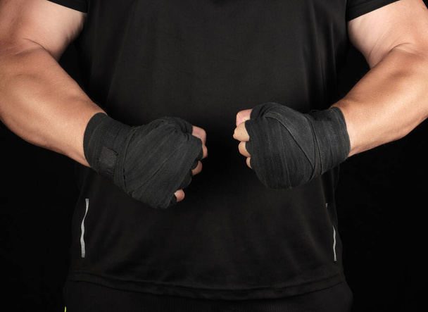 atleet in zwart uniform staat in een rek met gespannen spieren, zijn handen zijn verpakt in een zwart textiel verband, donkere achtergrond - Foto, afbeelding