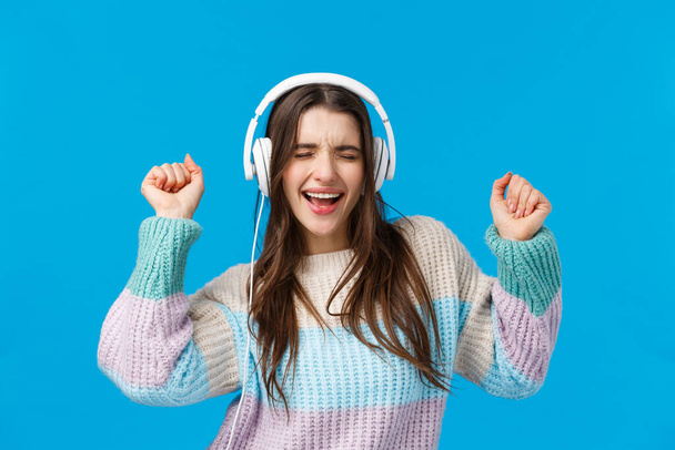 Rock n roll baby. Carefree emotive charismatic brunette woman enjoying listening music in new gift headphones, dancing raising hands up, close eyes and singing along favorite song, blue background - Photo, Image