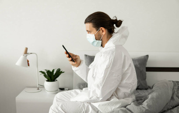Man in protective white suit and medical mask is using a phone at his home sitting on a bed because of coronavirus epidemic. Remote work during pandemic. Stay home during COVID-19 quarantine concept. - 写真・画像