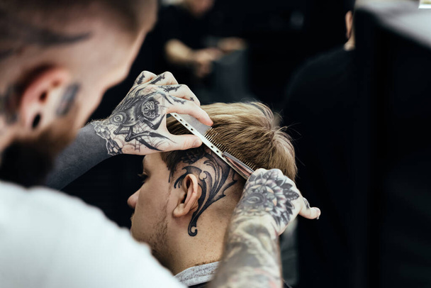 Getatoeëerde kapper knipt het haar van de klant met een schaar. Sluit maar af. Aantrekkelijke man is het krijgen van een modern kapsel in kapperszaak. - Foto, afbeelding
