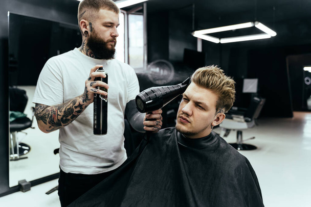 Barber makes hair styling with hair spray after haircut at the barber shop. Young handsome Caucasian man getting a haircut in a modern hairsalon. - Photo, Image