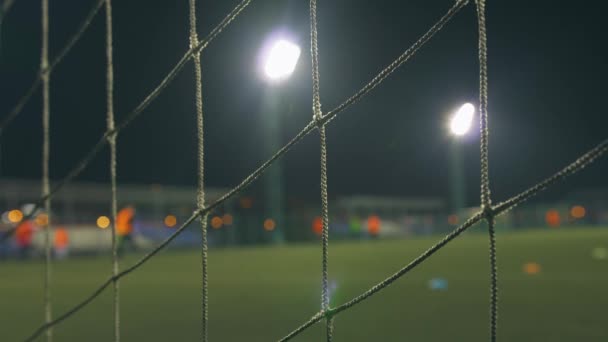 Abendliches Fußballspiel. Fußballer spielen auf dem Trainingsplatz, beleuchtet von Stadionlichtern. Blick durchs Tornetz. - Filmmaterial, Video