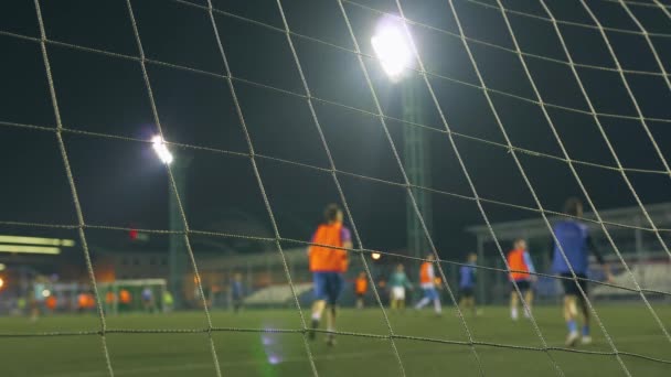 Partita di football serale. Giocatori di calcio giocano sul campo di allenamento, illuminato da luci dello stadio. Vista attraverso la rete degli obiettivi
. - Filmati, video