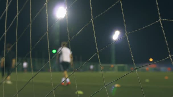 Avond voetbalwedstrijd. Voetbalspelers spelen op het trainingsveld, verlicht door stadionlichten. Zicht door doelnet. - Video