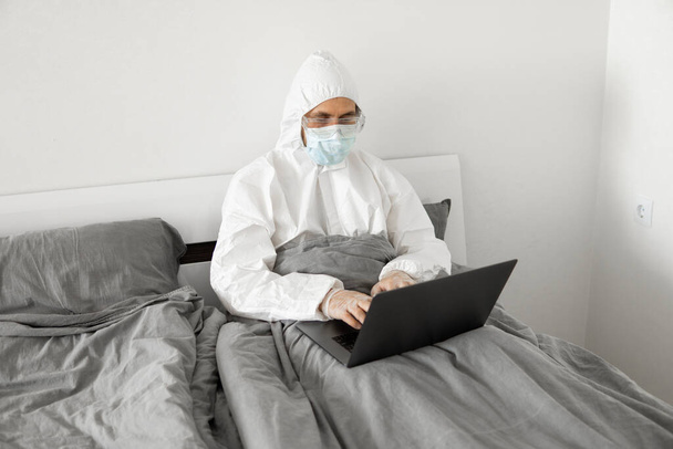 Man in protective white suit and medical mask is working from home in a bed with laptop and speaking with his friend or business partners via webcam. Coronavirus epidemic. Remote work during pandemic. - Foto, Imagem