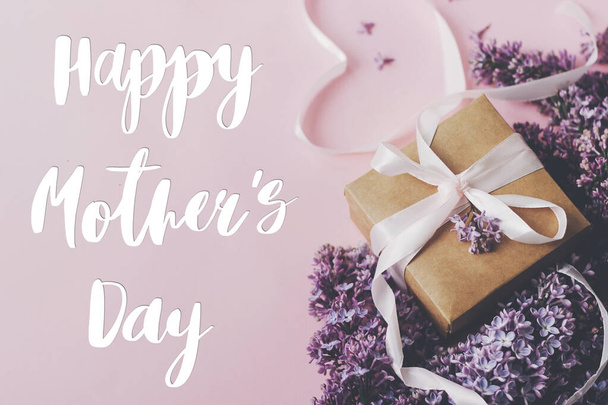 Happy Mother 's day text and lilac flowers, gift box and heart ribbon on pink paper, stylish flat lay. Фиолетовый букет сиреневых цветов с коробкой подарков. Открытки на день матери
 - Фото, изображение