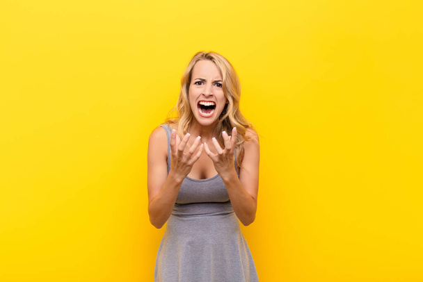 young blonde woman looking desperate and frustrated, stressed, unhappy and annoyed, shouting and screaming against orange wall - Foto, imagen