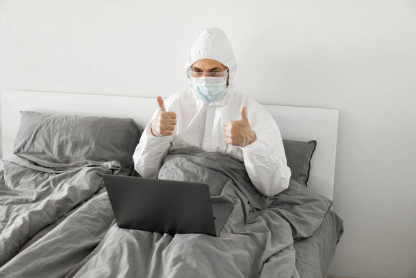 Man in protective white suit and medical mask is working from home in a bed with laptop and speaking with his friend or business partners via webcam. Coronavirus epidemic. Remote work during pandemic. - Foto, Imagen