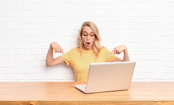 young blonde woman with open mouth pointing downwards with both hands, looking shocked, amazed and surprised using a laptop - Фото, зображення