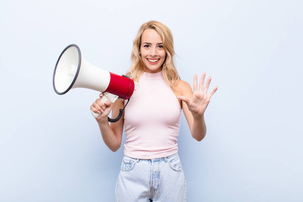 young blonde woman smiling and looking friendly, showing number five or fifth with hand forward, counting down with a megaphone - Fotoğraf, Görsel
