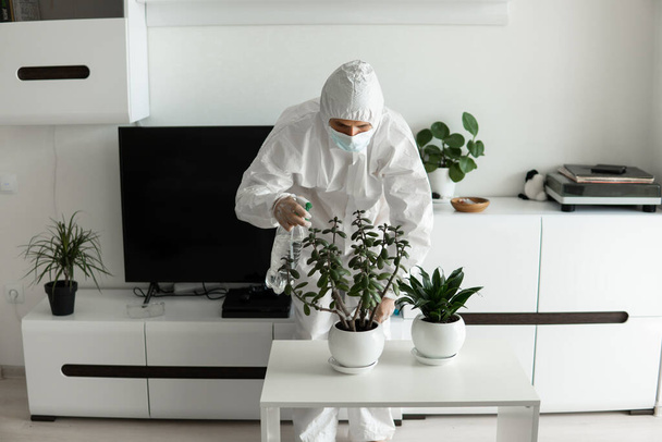 Muž v ochranném obleku, lékařské masce a gumových rukavicích pečuje o rostliny s postřikovačem doma v obývacím pokoji, zatímco koronavirus karanténa COVID-19. - Fotografie, Obrázek