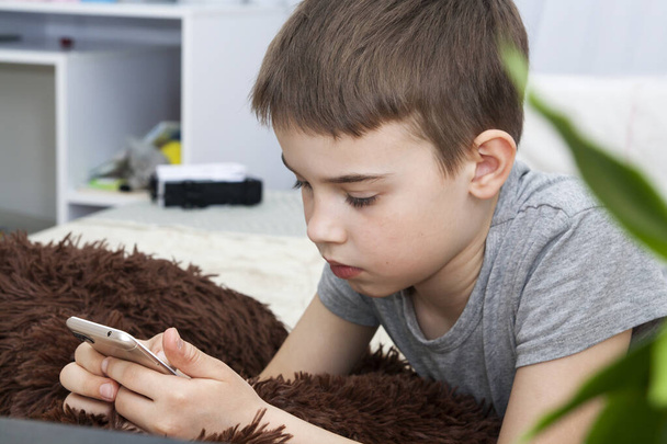 8-9 yaşlarında bir çocuk elinde telefonuyla yatakta yatıyor. Evde eğitim. Karantina koronavirüsü evde çocukları izole ediyor. - Fotoğraf, Görsel
