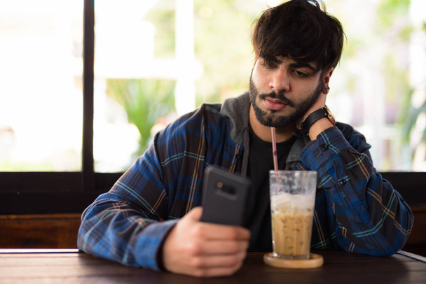 Portrait of young handsome bearded Indian hipster man relaxing at the coffee shop - Foto, imagen