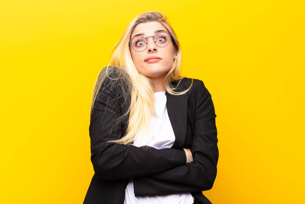young pretty blonde woman shrugging, feeling confused and uncertain, doubting with arms crossed and puzzled look against yellow wall - Photo, Image