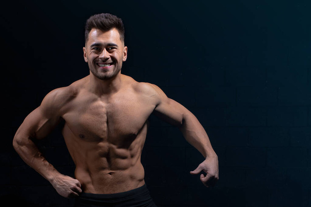 male athlete shows body and muscles on dark wall background in gym, sexy guy bodybuilder, sports lifestyle, home sport - Fotografie, Obrázek