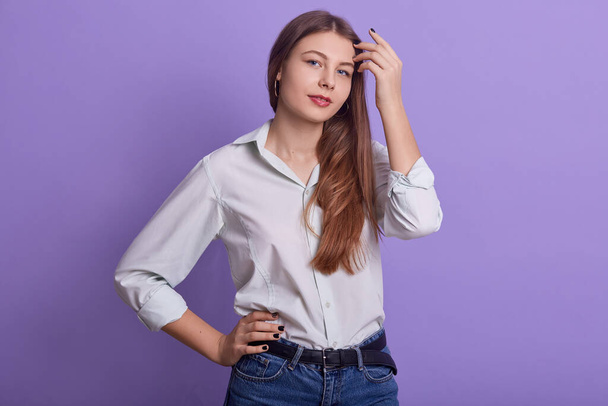 Horizontal portrait of beautiful sweet young female touching her hair, keeping hand on waist, being in good mood, wearing jeans and light shirt, posing isolated over lilac background in studio. - Φωτογραφία, εικόνα