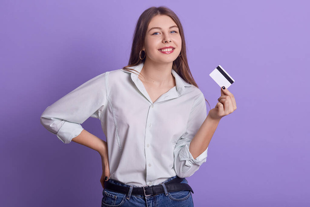 Portrait of funny attractive young female holding credit card in one hand, smiling sincerely, looking directly at camera, wearing jeans and light shirt, keeping hand on waist. Expenses concept. - 写真・画像