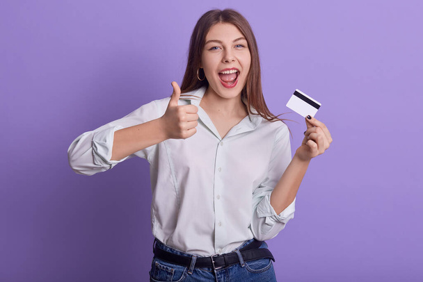 Picture of emotional sweet energetic young woman holding credit card, making gesture, thumb up, showing approval, opening mouth widely, wearing light shirt and jeans. People and money concept. - Zdjęcie, obraz
