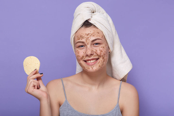 Attractive young woman with her hair wrapped in white towel, girl with perfect skin cleansing her face, using coffee scrub, posing isolated over lilac background, holding cotton sponge in hand. - Foto, Imagem