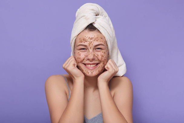 Indoor shot of funny young girl with white towel on her head, lady with coffee scrub on her face, charming woman using cleansing mask, keeping fists under chin, looking at camera. cosmetology concept. - Photo, Image