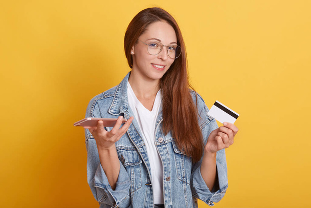 Horizontal picture of cheerful cute good looking lady having delighted facial expression, holding credit card and mobile phone, using new technologies, buying online. Women and shopping concept. - Фото, изображение