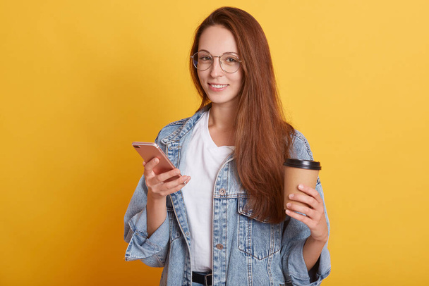 Picture of cheerful good looking young female standing isolated over yellow background in studio, holding paper cup with hot drink and smartphone, having delighted facial expression. Emotions concept. - Fotografie, Obrázek