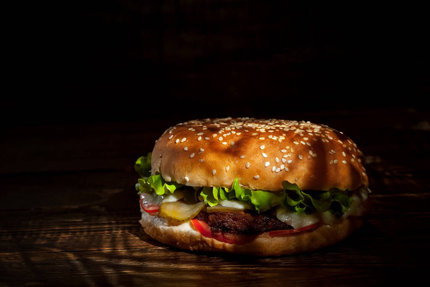 Burger on a dark wooden board. Cheeseburger with cutlet and herbs on black background. Food with contrasting dramatic light. Bun closeup. - Photo, image