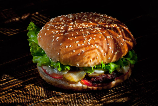 Burger on a dark wooden board. Cheeseburger with cutlet and herbs on black background. Food with contrasting dramatic light. Bun closeup. - Photo, Image