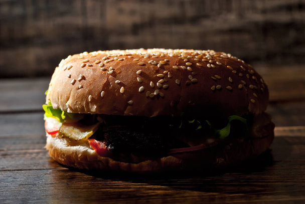 Burger on a dark wooden board. Cheeseburger with cutlet and herbs on black background. Food with contrasting dramatic light. Bun closeup. - Photo, Image