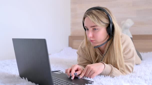 Young girl freelancer works at a laptop at home in her bed. Young woman wears headset conference calling on laptop talks with online teacher studying. Distance learning. Work from home concept. - Video