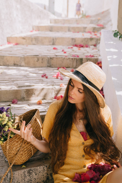 girl in a yellow dress and hat with long brown hair, is sitting on the steps, on the background of green flowering cactus and pink bougainvillea flowers, in summer in Mediterranean, on vacation - Foto, Imagen