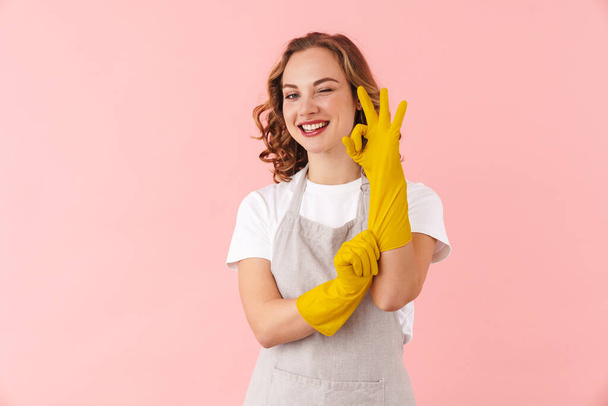 Image of a positive cheery young woman housewife in gloves isolated over pink wall background showing okay gesture. - Foto, imagen