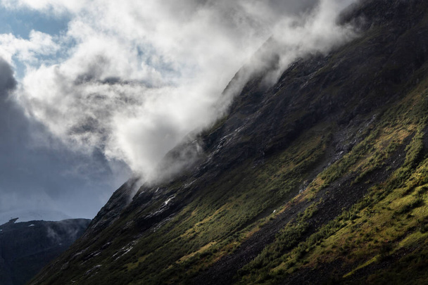 Dağ zirvesi bulutlarla kaplı ve yeşil tepede parlak güneş ışınları var. Norveç Avrupa 'sında Troller Yolu Trollstigen Yolu manzarası - Fotoğraf, Görsel
