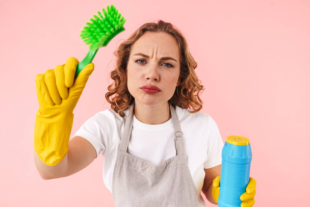 Image of serious concentrated woman housewife isolated over pink wall background in gloves holding cleanser and brush. - Фото, изображение