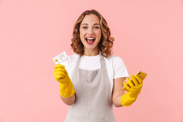 Image of a happy excited young housewife woman in gloves holding mobile phone and credit card isolated over pink wall background. - Foto, Imagen