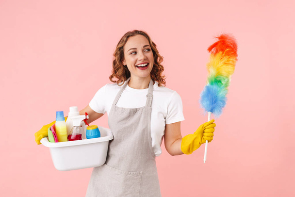 Image of optimistic young housewife woman in gloves holding cleanser bottles and colorful duster isolated over pink wall background. - Photo, image