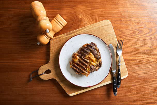 top view of fresh grilled steak on plate on cutting board with cutlery and salt and pepper mills on wooden table - Foto, imagen