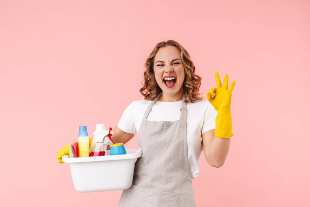 Imagen de una feliz mujer joven emocionada ama de casa con guantes aislados sobre fondo de pared rosa sosteniendo limpiadores que muestran un gesto bien
. - Foto, imagen