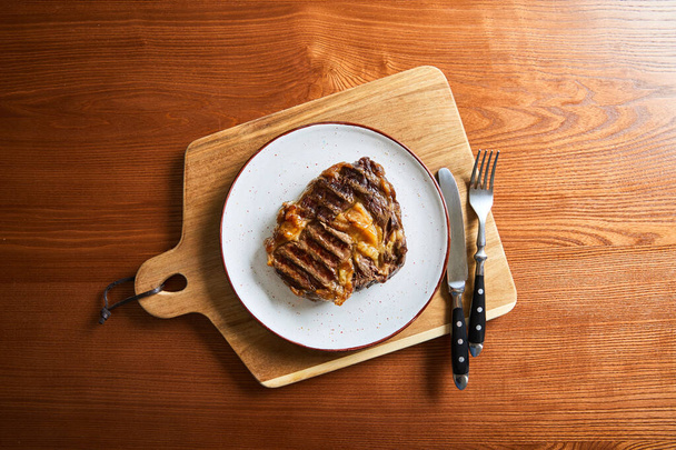 ylhäältä näkymä tuoretta grillattua pihviä lautasella leikkuulauta ruokailuvälineet puinen pöytä
 - Valokuva, kuva