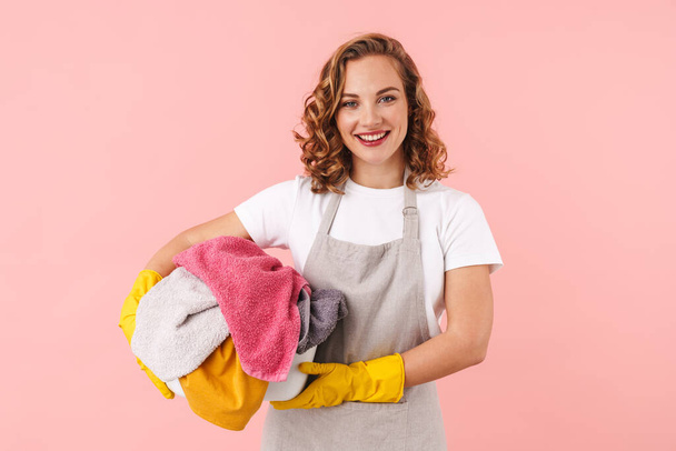 Picture of a cheery happy young woman housewife in gloves isolated over pink wall background holding laundry. - Zdjęcie, obraz