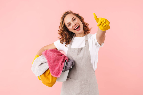 Picture of a positive happy young woman housewife in gloves isolated over pink wall background holding laundry showing thumbs up. - Foto, afbeelding