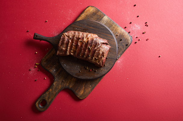 top view of tasty grilled steak served on wooden boards on red background with pepper and salt - Foto, Imagem