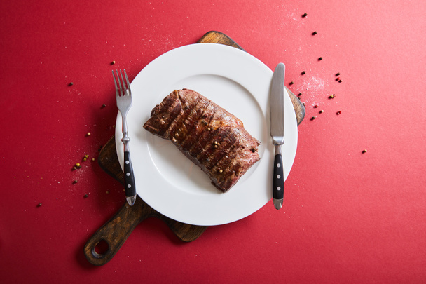 top view of tasty grilled steak served on plate on wooden board on red background with pepper and salt - Valokuva, kuva