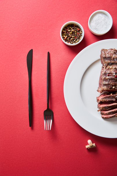vue de dessus de steak grillé savoureux servi dans une assiette avec couverts et sel et poivre dans des bols sur fond rouge
 - Photo, image