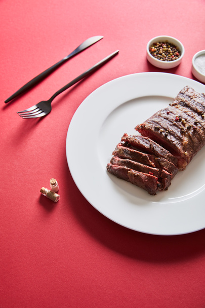 sabroso bistec a la parrilla servido en el plato con cubiertos y sal y pimienta en cuencos sobre fondo rojo
 - Foto, Imagen