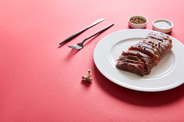 leckeres Steak vom Grill serviert auf Teller mit Besteck und Salz und Pfeffer in Schüsseln auf rotem Hintergrund - Foto, Bild