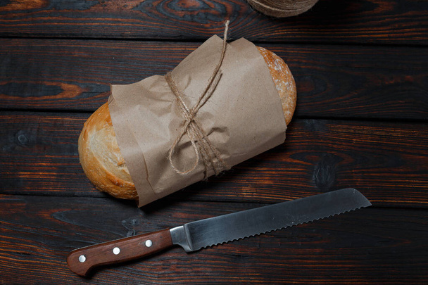 Επίπεδη στρώση φρεσκοψημένου ψωμιού και μαχαιριού σε χαρτί χειροτεχνίας σε ρουστίκ ξύλινη σανίδα σε σκούρο ξύλινο τραπέζι, πάνω όψη, επιλεκτική εστίαση. - Φωτογραφία, εικόνα