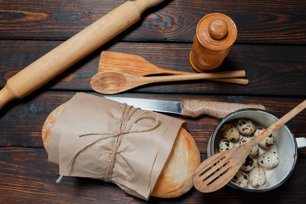 Brot in Bastelpapier eingewickelt, Wachteleier liegen in einem Becher, ein Messer, hölzerne Küchenutensilien liegen auf einem dunklen Holztisch. Draufsicht, Kopierraum. Selektiver Fokus. - Foto, Bild