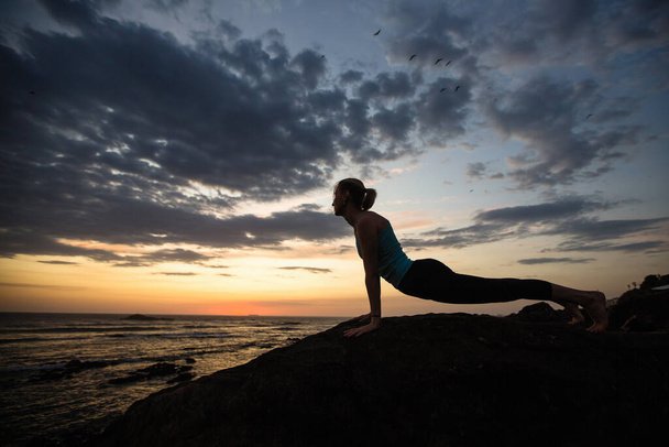 Γιόγκα γυναίκα κάνει ασκήσεις στην ακτή του ωκεανού στο λυκόφως. Πορτογαλία. - Φωτογραφία, εικόνα
